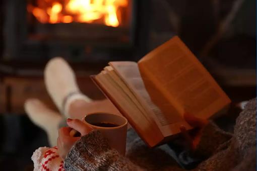 A woman relaxes in front of a fire with a book and a hot drink.